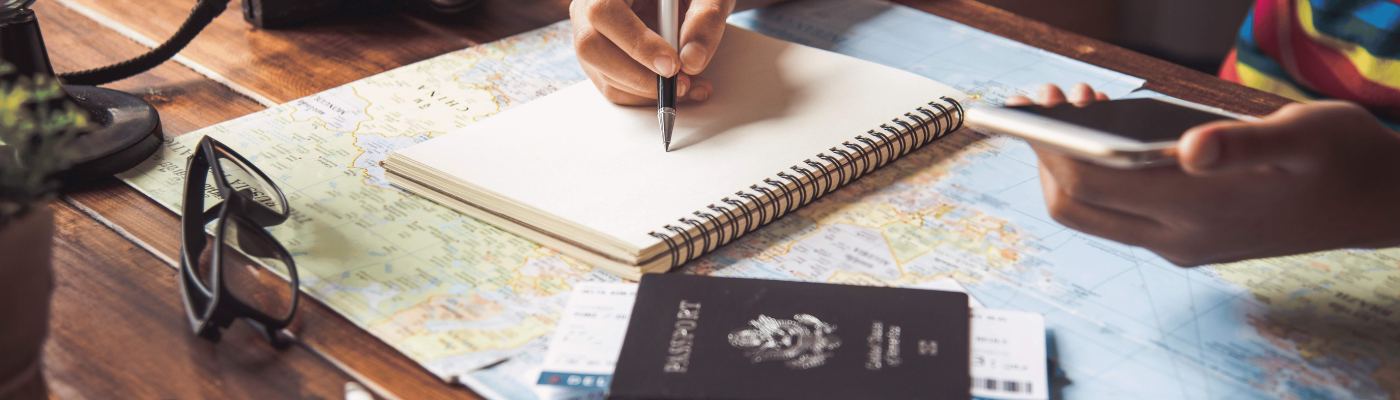A person writes in a notebook on a map-covered table, holding a phone. Nearby are passports, boarding passes, a camera, a toy plane, and a globe, suggesting travel planning.