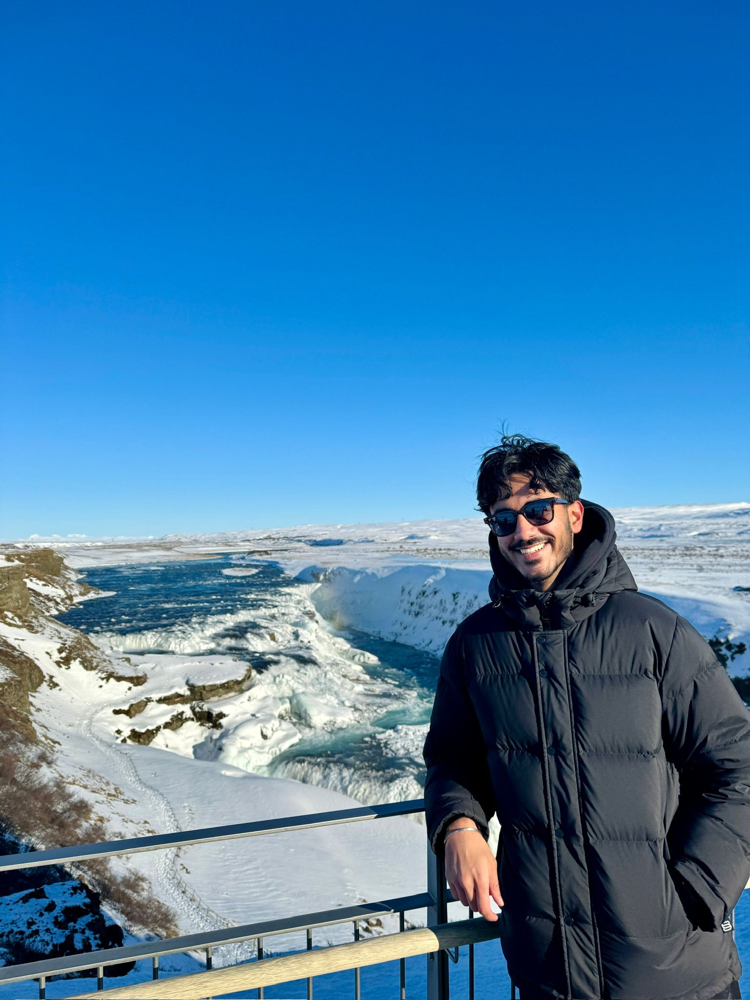 A person wearing a black coat and sunglasses smiles while standing by a railing with a snowy landscape and a large waterfall under a clear blue sky in the background.