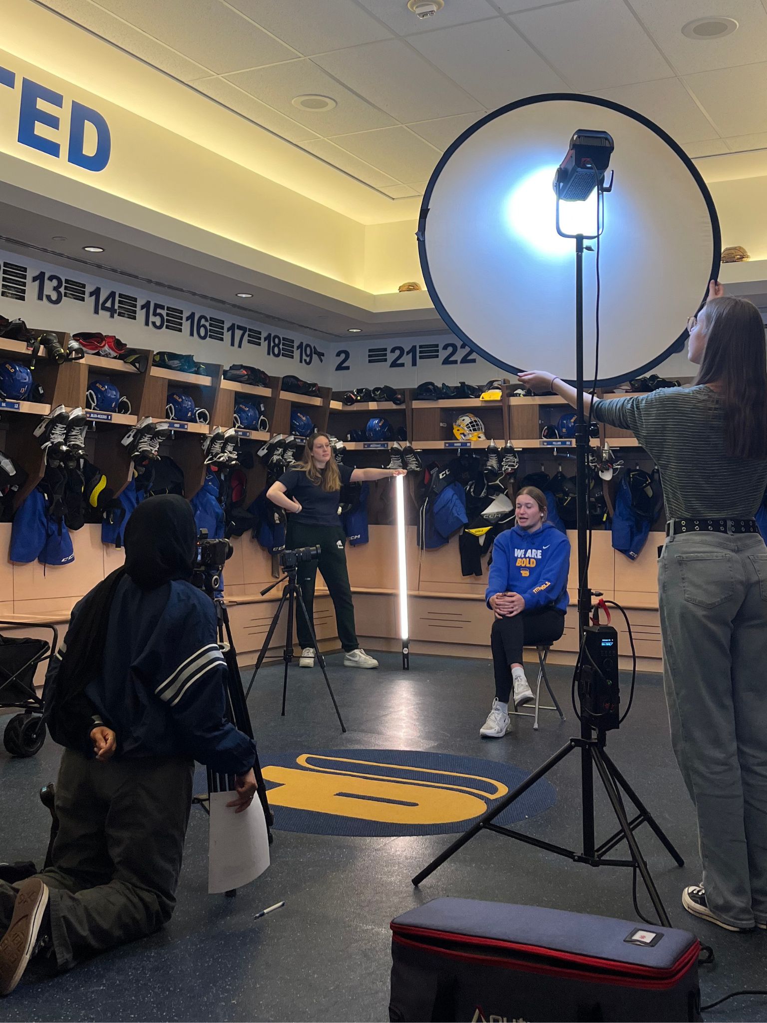 A woman sits smiling for a photoshoot in a sports locker room, surrounded by three crew members adjusting lights and equipment. Blue and yellow uniforms and helmets hang in the background above numbered lockers