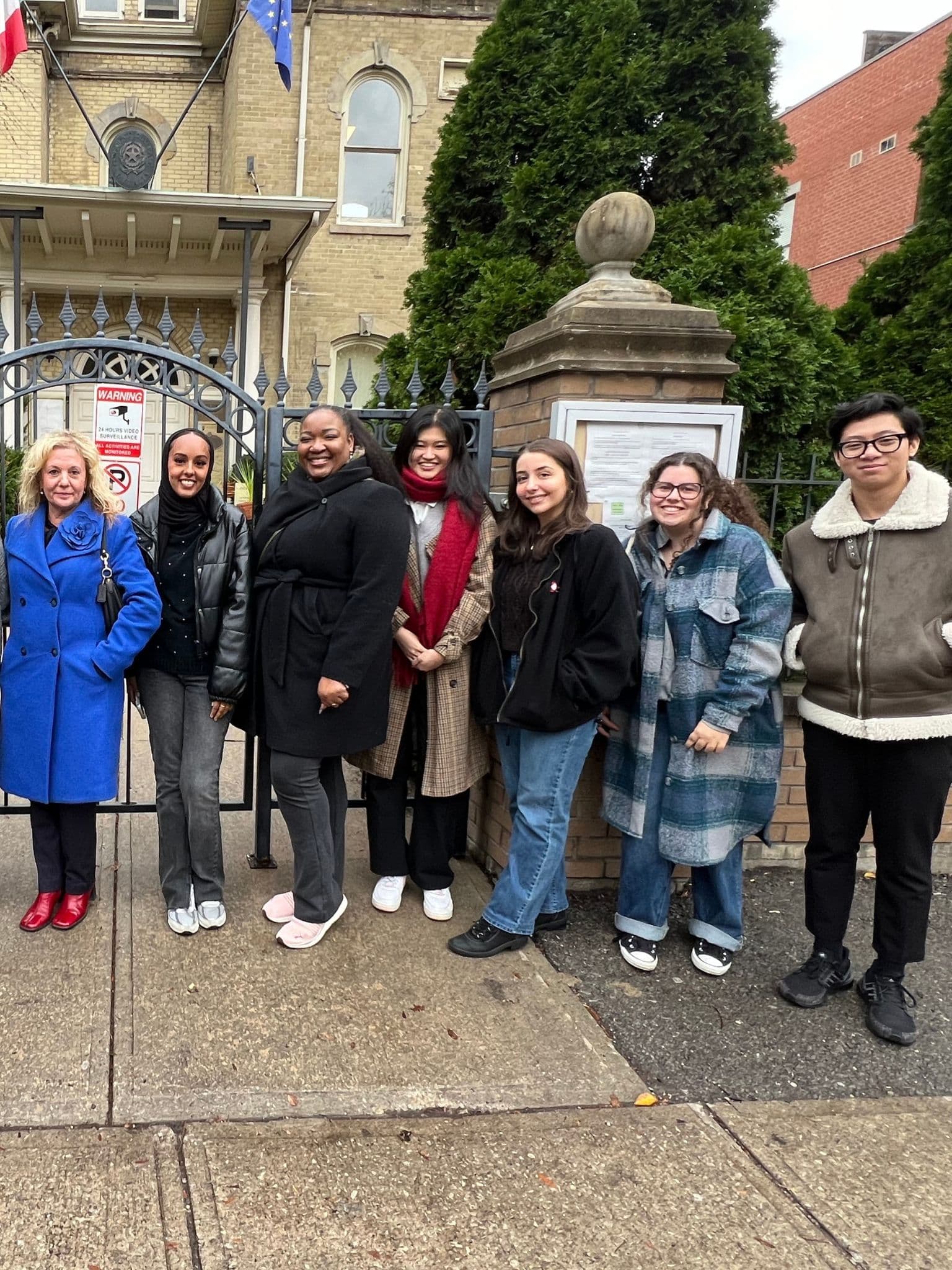 Seven people stand smiling in a row on a sidewalk in front of a historic stone building and iron gate, dressed in warm coats and casual clothes on a cloudy day.