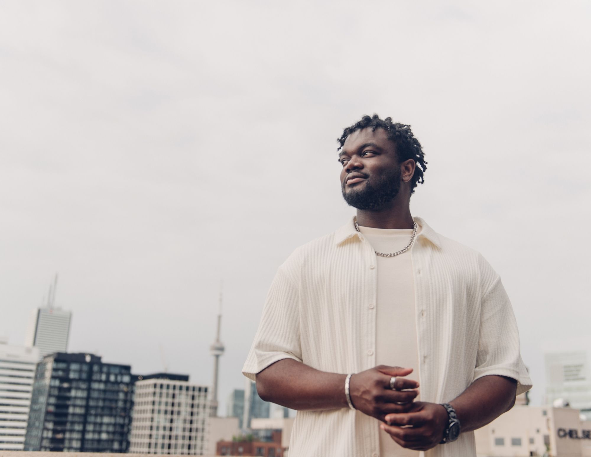 A man stands outdoors in front of the toronto skyline, wearing a light-colored shirt and looking to the side with a slight smile. The sky is overcast and tall buildings are visible in the background.