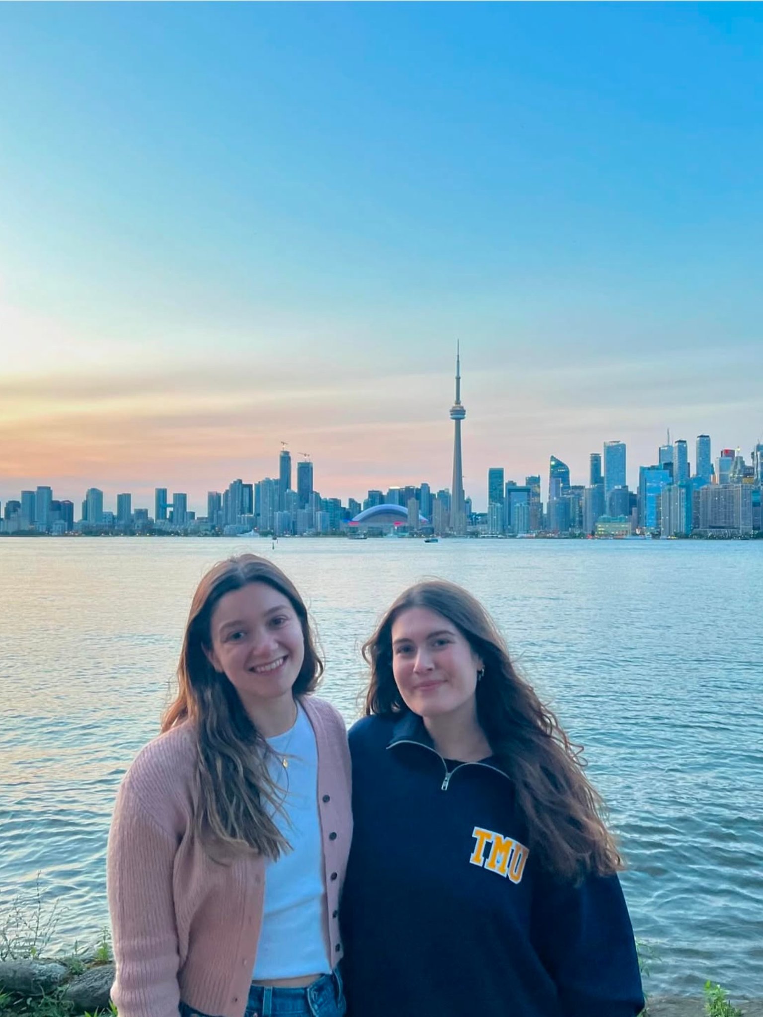 Two young women stand smiling by the waterfront at sunset, with the Toronto city skyline and CN Tower visible in the background under a clear blue and orange sky.