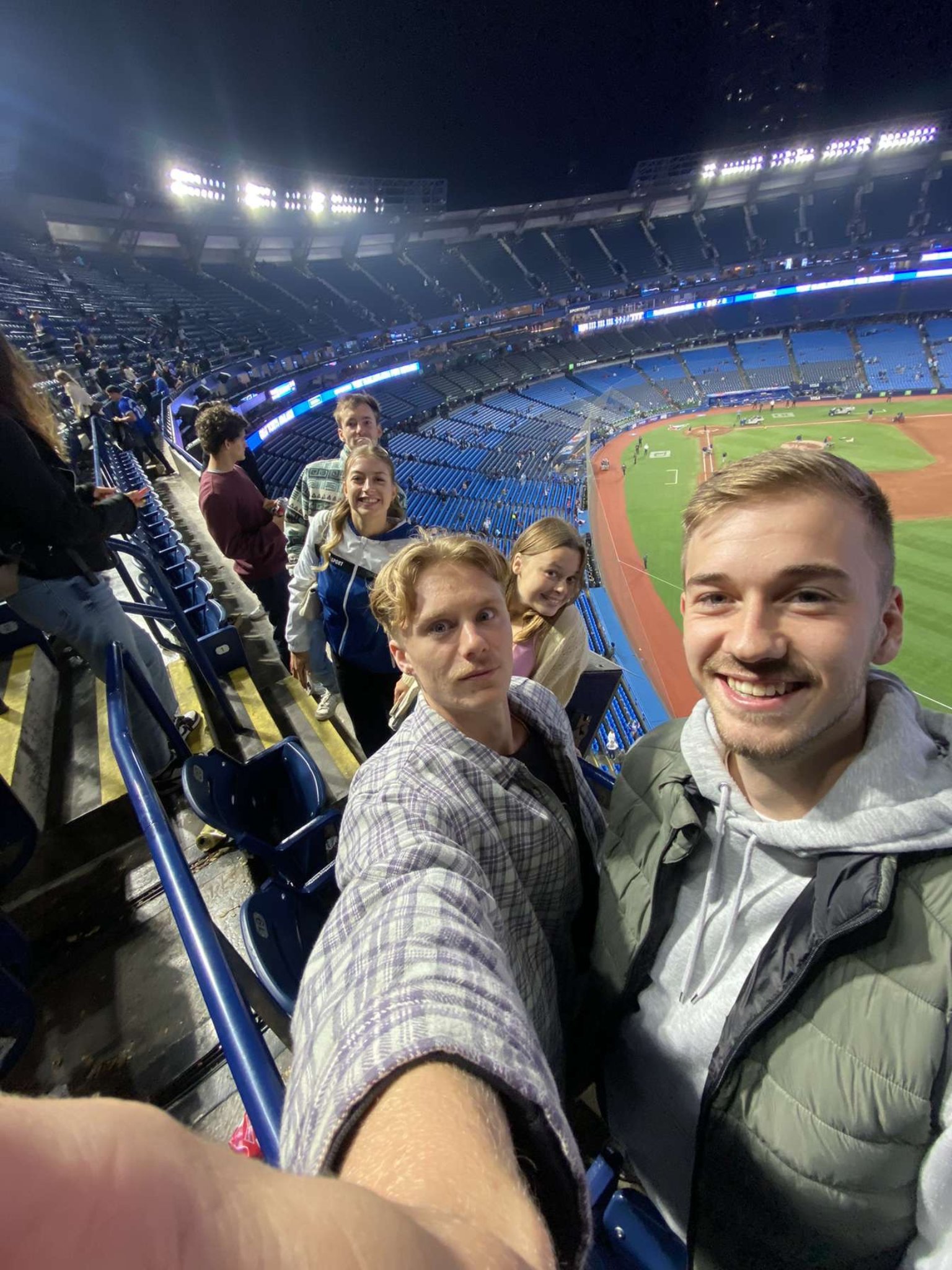 A group of five young adults taking a selfie and smiling in rogers centre, with blue seats and a lit field visible in the background.