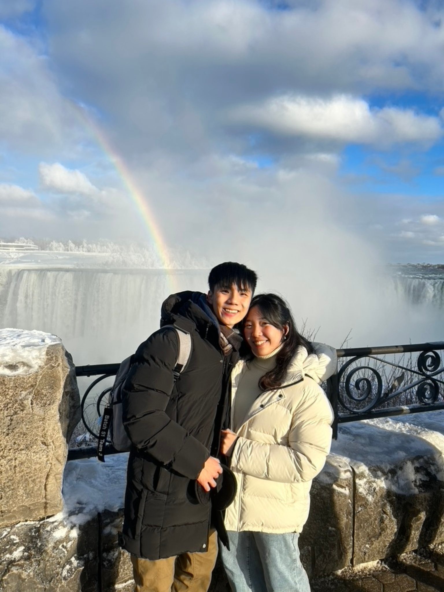 two young students in winter coats smiles in front of a misty waterfall with a rainbow in the background. Snow covers the stone railing and ground, and the sky is partly cloudy.