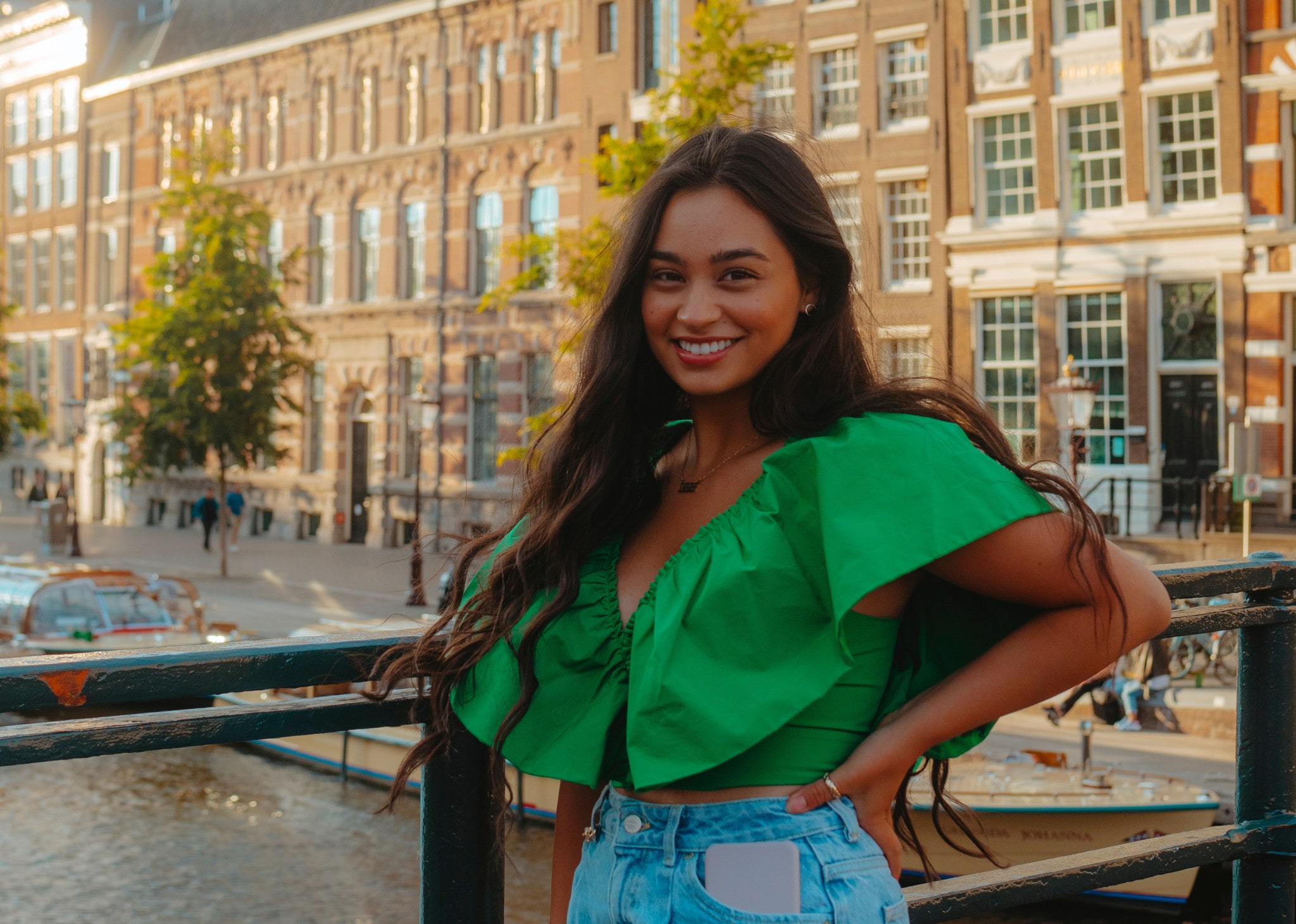 Smiling student standing on a bridge over a canal in Amsterdam, wearing a bright green top and jeans, with historic brick buildings in the background.
