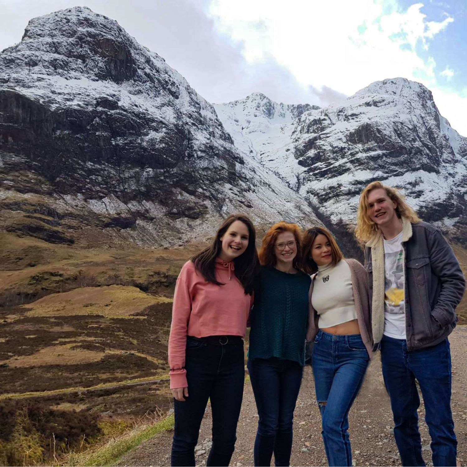 Four students standing together on a mountain road, smiling with snow-covered peaks in the background.