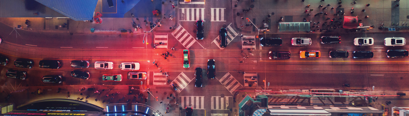 Aerial view of a busy city intersection at night with cars, crosswalks, and people walking under colorful streetlights.