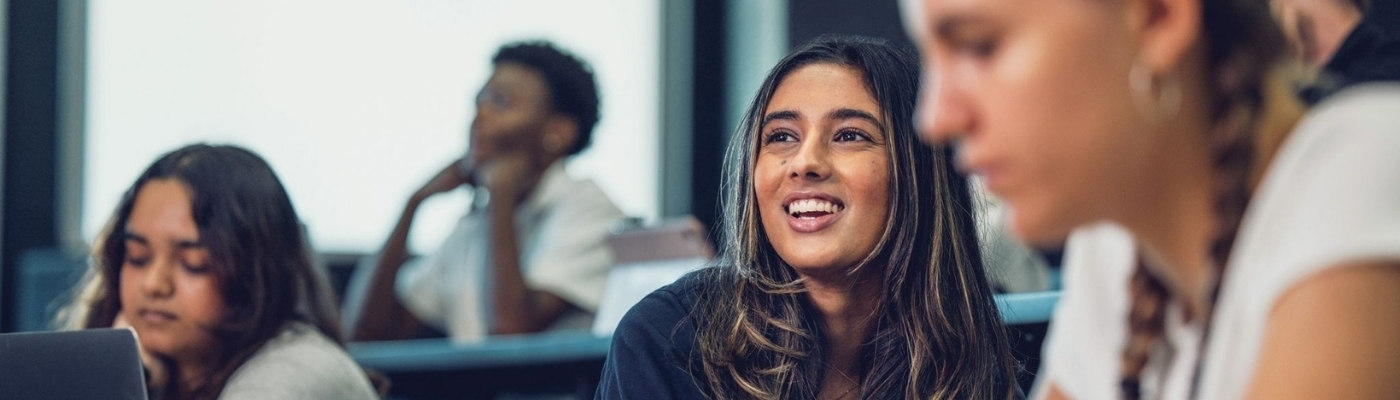 A group of students sit in a classroom; one student in the center smiles and looks engaged, while others are focused on their devices or notes. The atmosphere appears relaxed and attentive.
