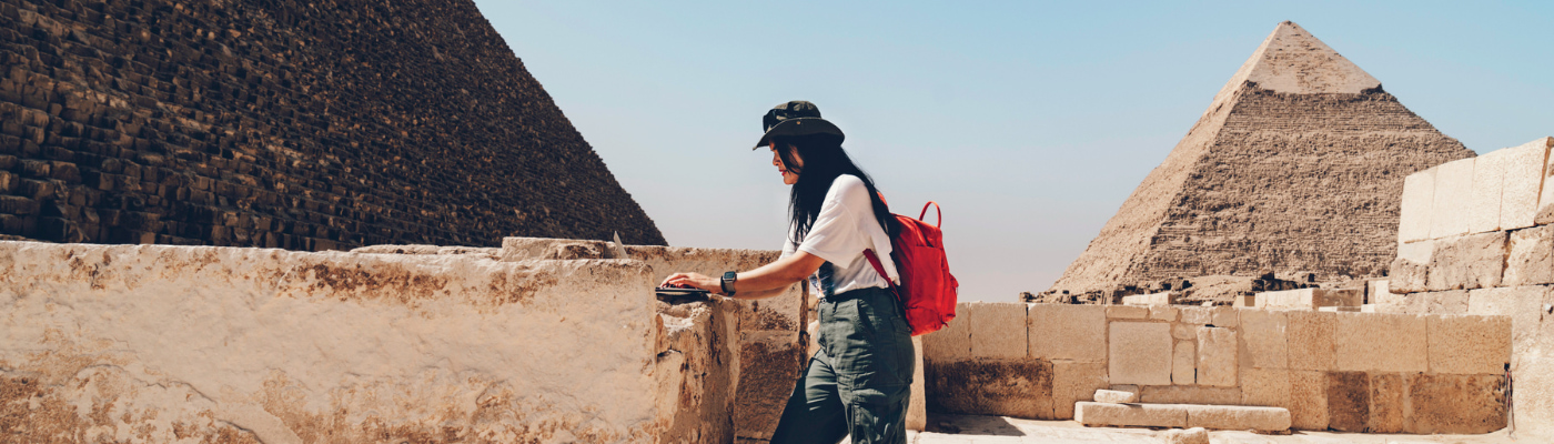 Person wearing a hat and red backpack standing near stone ruins, with two large pyramids in the background under a clear blue sky.