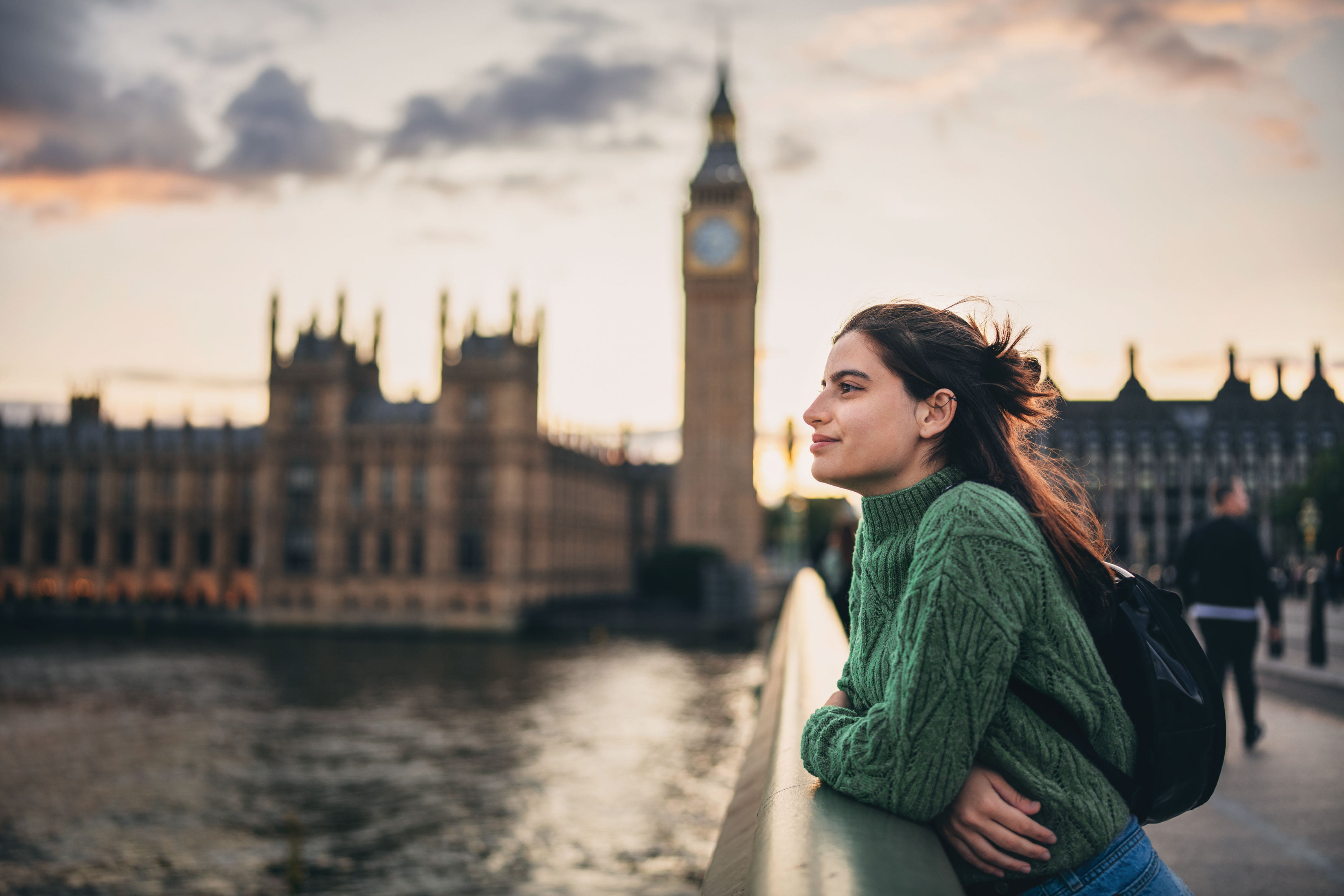Student in a green sweater leaning on a bridge railing, smiling while overlooking the River Thames with Big Ben and the Houses of Parliament in the background at sunset in London.