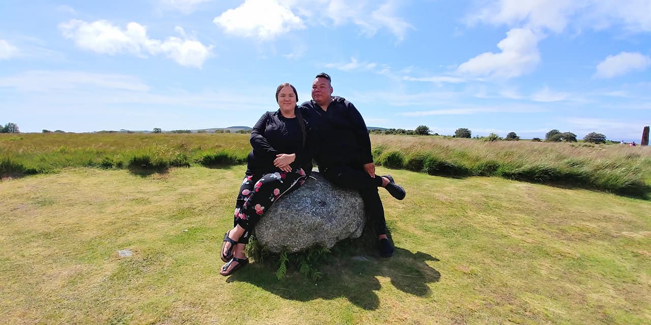 The Indigenous hosts of the series sit smiling next to each other on a boulder in the middle of a green field. In the background are tall grasses and blue sky with clouds. 