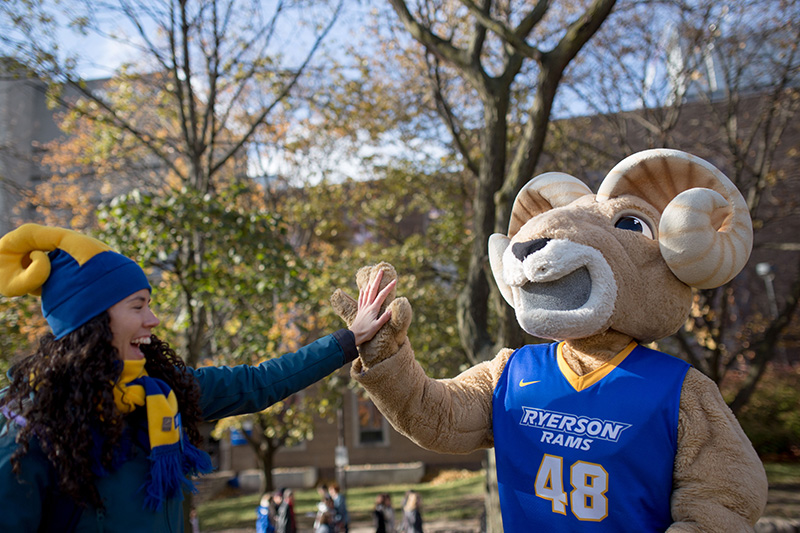 A female student wearing a Ryerson Varsity jacket is interacting with Ryerson's mascot, Eggy the Ram 
