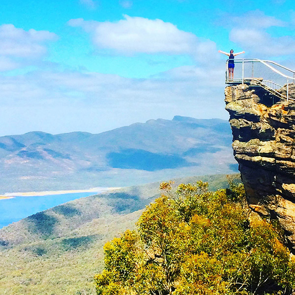 Toronto Metropolitan University student on exchange poses on cliff's edge with mountain range in the background on a sunny day.