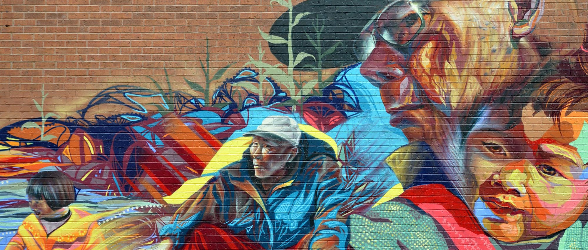 A mural painted on a brick wall depicting the journey, possibly an immigrant man, from as a child to an elder. The child is painted in a mix of brown, yellow, blue colours, and the man is depicted in a dark blue, grey and navy colours with a stern, composed face.