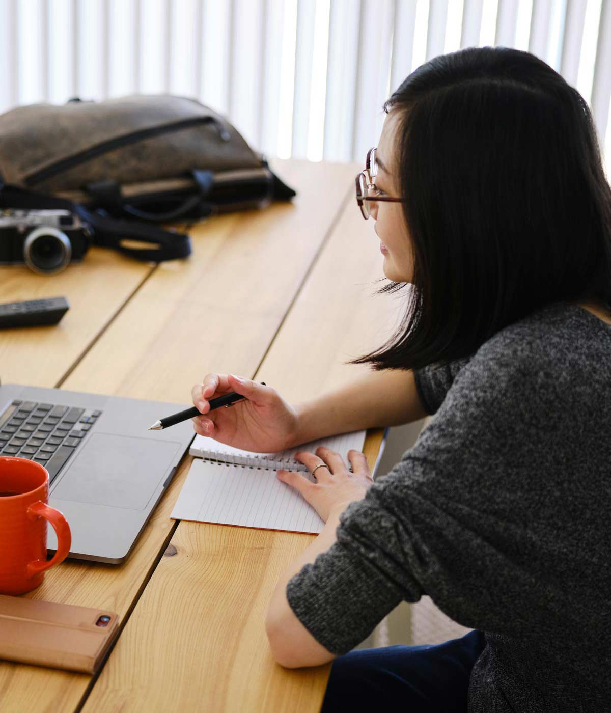 A woman with glasses works on a laptop at a wooden table, holding a pen over a notebook. Nearby are an orange mug, a camera, and a backpack.