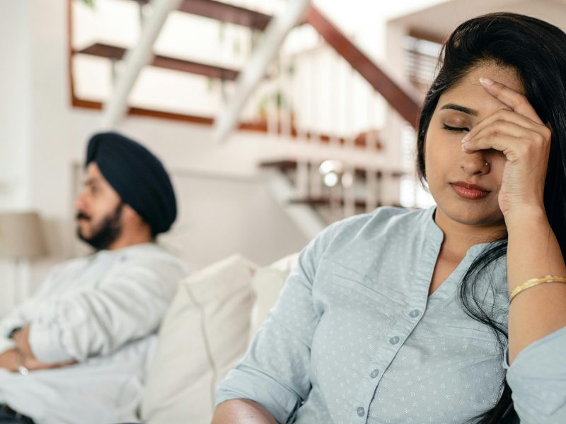 Woman sitting on couch in home beside man with crossed arms looking upset with head in hand
