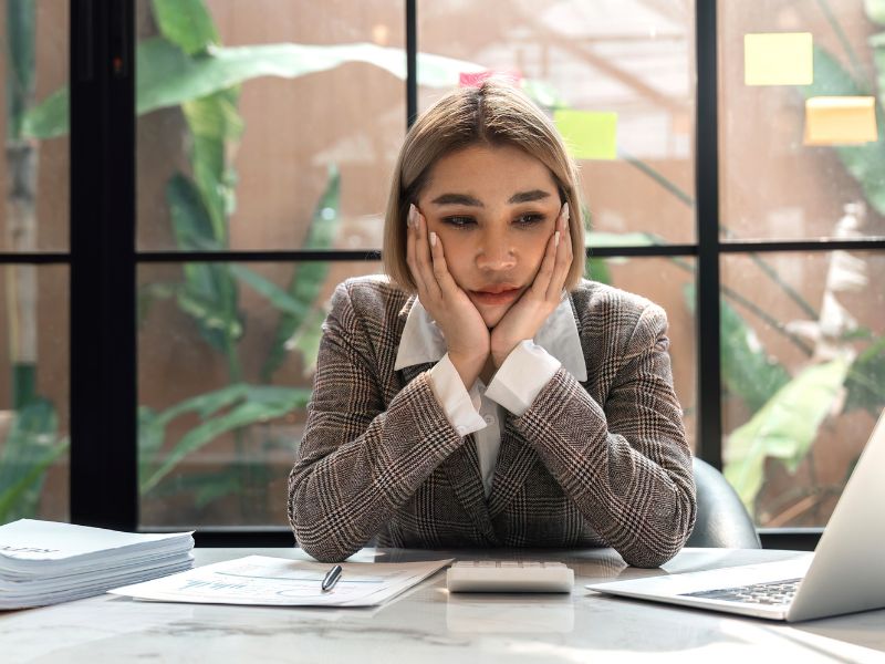 A woman in a business suit sits at a desk, resting her hands on her face, appearing deep in thought