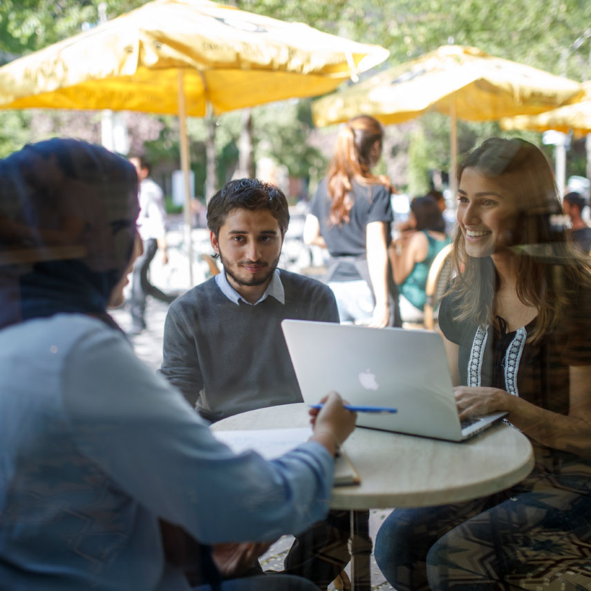 Three students sitting at Balzac's outdoor patio table. Student with notebook and pen sits across from student with laptop.