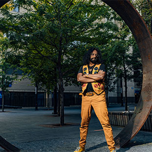 South Asian man with long black hair, glasses and orange vest with embroidered flowers standing with arms crossed in front of steel Ring sculpture on TMU campus