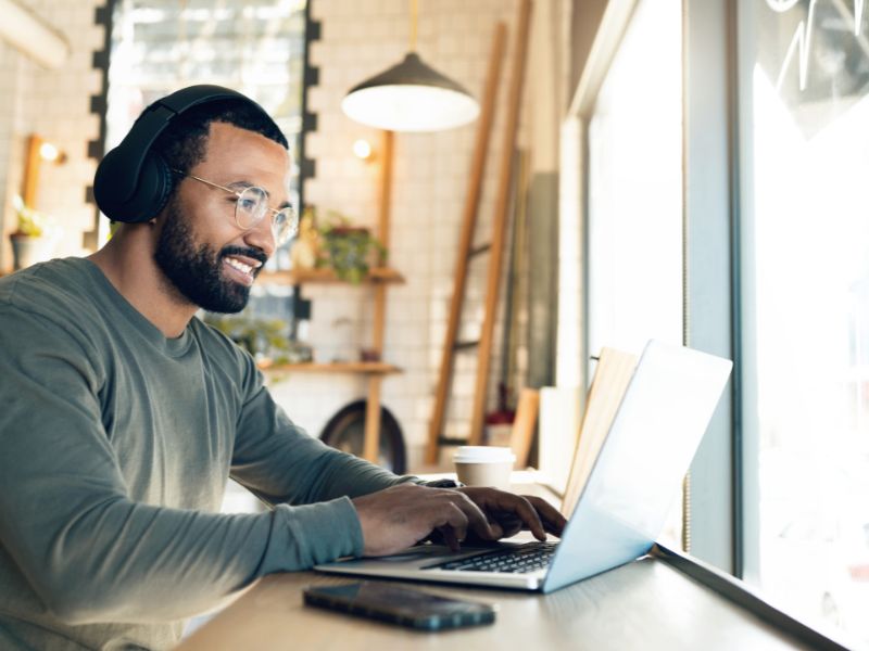 Male graduate student sitting in cafe typing on open laptop on desk with headphones on