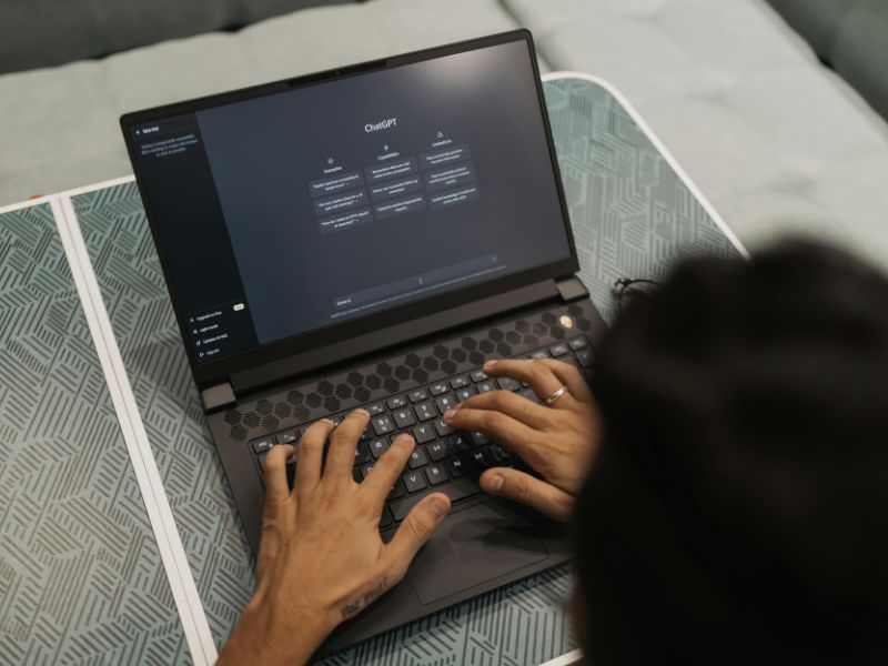 A person typing on a laptop displaying ChatGPT in a dimly-lit room. The laptop is on a patterned table, suggesting a focused, tech-focused atmosphere