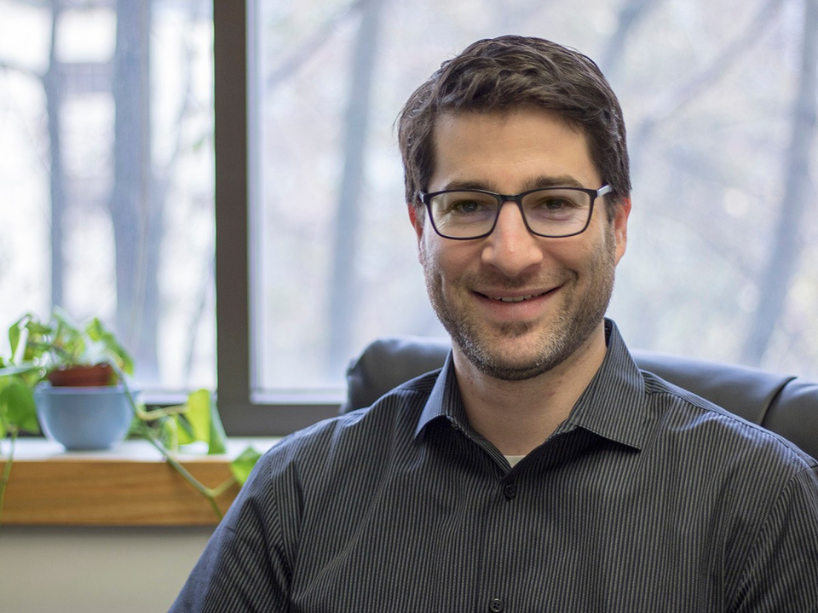 Dr. Seth Dworkin sits at a desk in front of a window wearing a dark shirt and glasses smiling at the camera