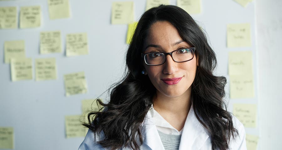 A woman in a white lab coats stands in front of a wall of sticky notes