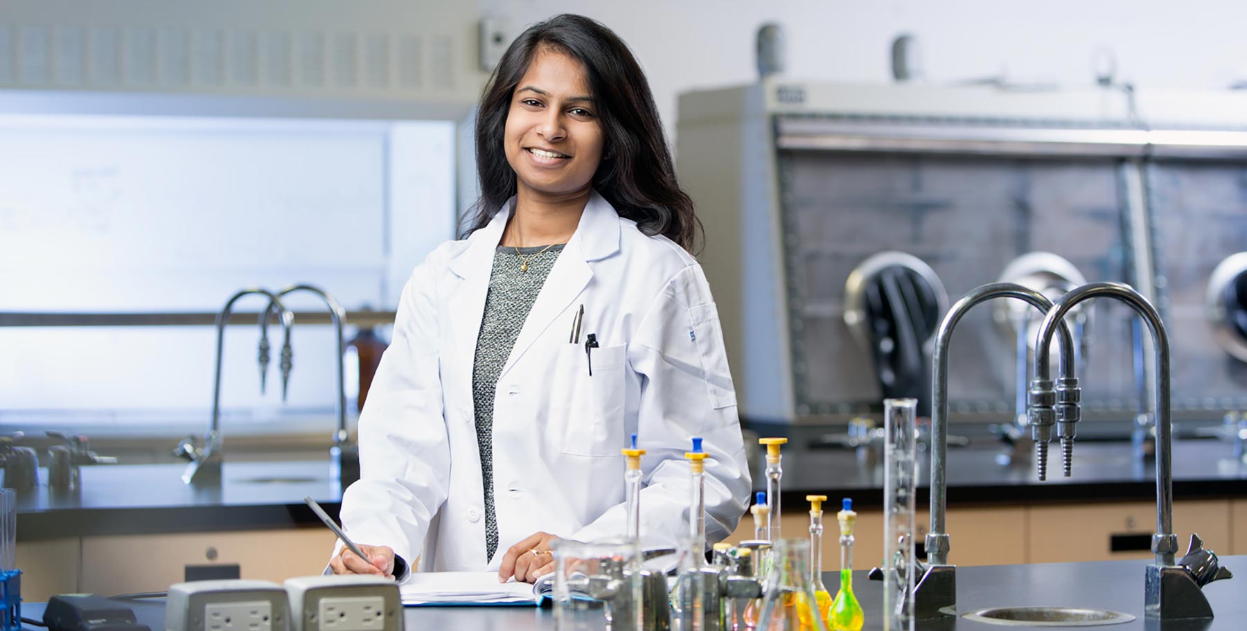 A woman in a white lab coat stands behind her lab workstation