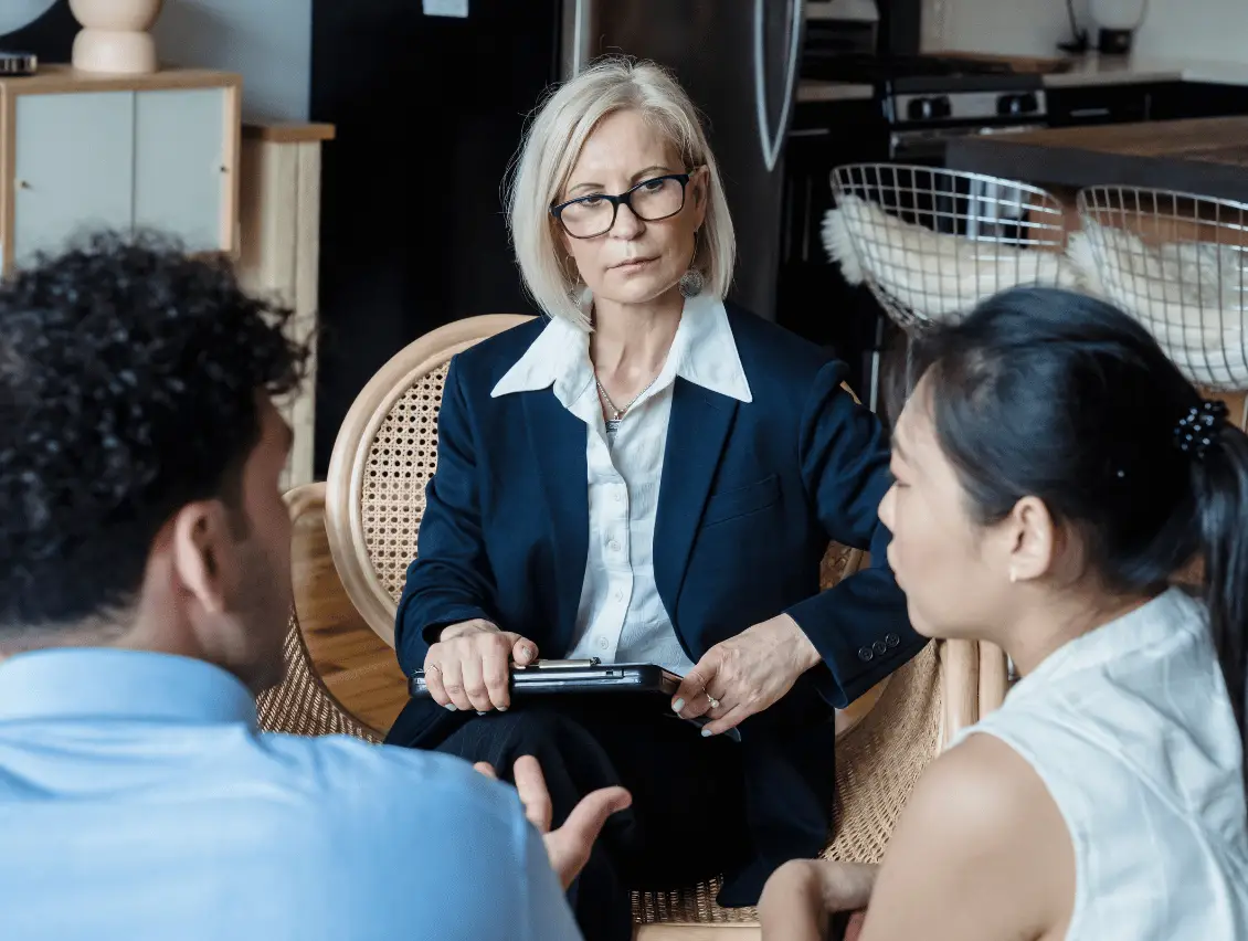 Older woman in glasses and navy blazer holds a tablet while speaking with two younger colleagues in a bright, professional office