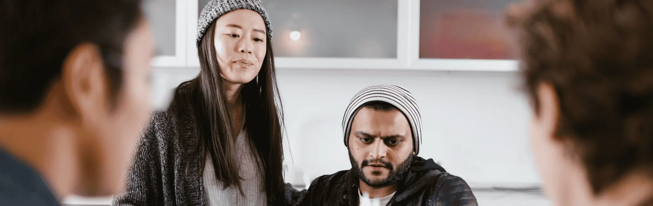 Female graduate student in a knit hat stands beside a concerned seated male graduate student, while two blurred people listen in the foreground