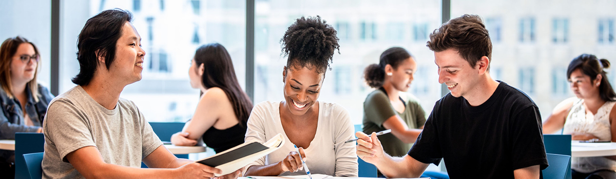 A group of students sits around a table, smiling and collaborating on coursework while other students study in the background.