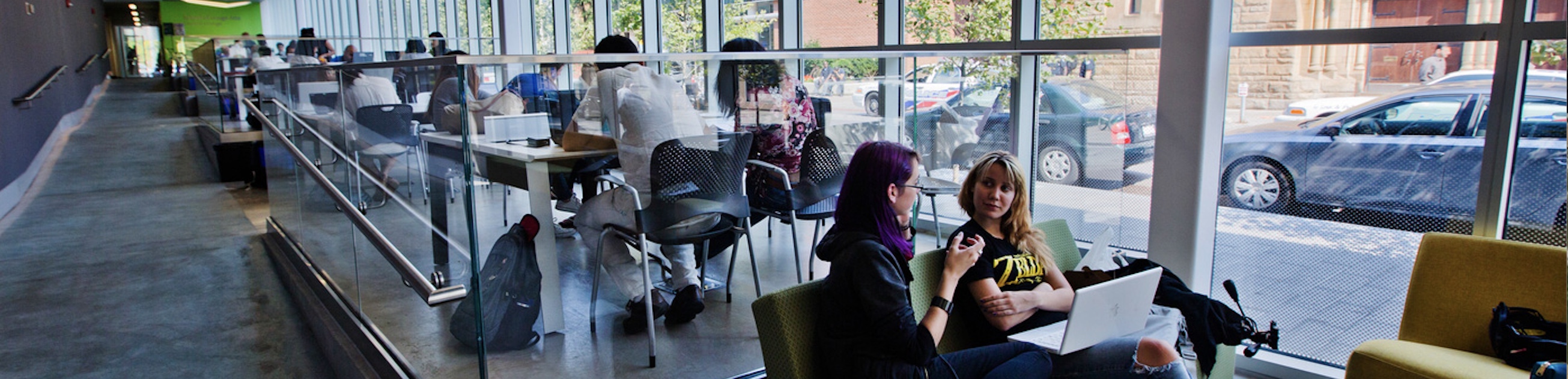 Two students talking in a modern, glass-walled university study space with other students working in the background.