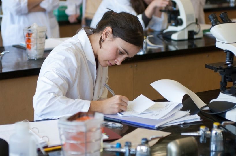 A student in white lab coat writes notes in a lab.