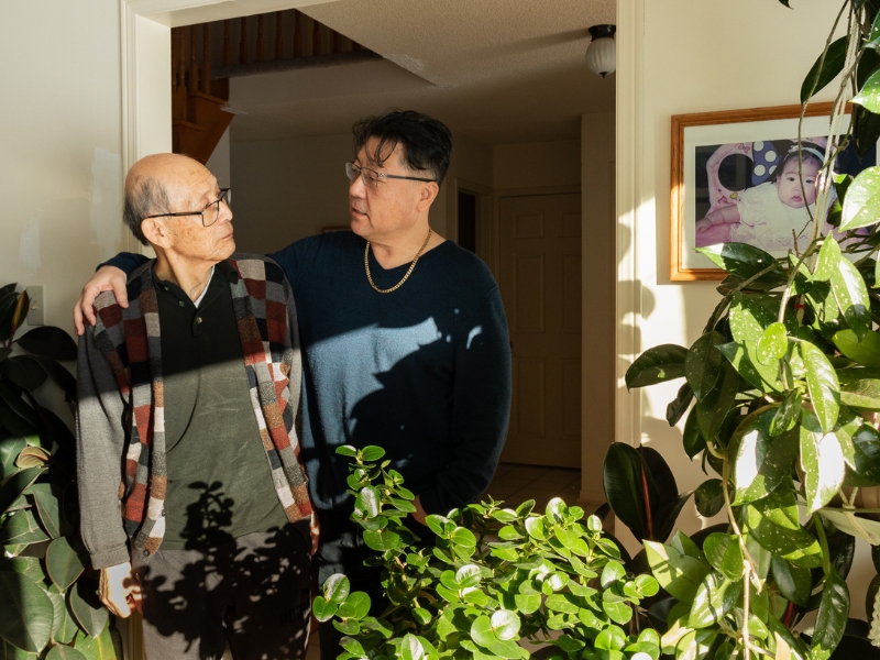 Elderly man in a patterned cardigan and younger man in a dark sweater share a warm moment, surrounded by lush indoor plants and a framed baby photo.