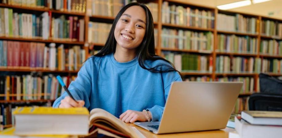 Female Asian student sitting at desk in library with open laptop and smiling.