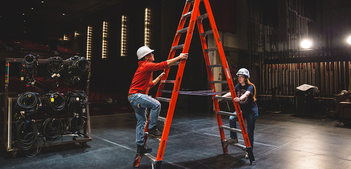 Two people using a tall ladder on stage at the TMU theatre. Both wearing safety gear, one person is climbing, the other person is holding for support.