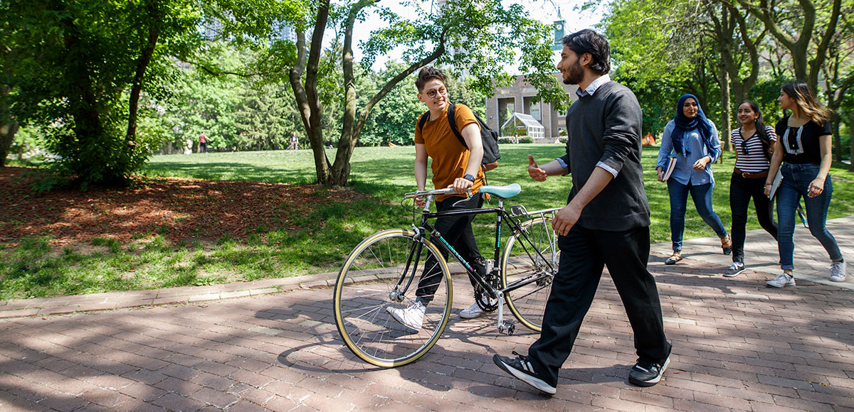 Bright spring day on in the TMU Quad, students walk on a path, one is pushing a bike.