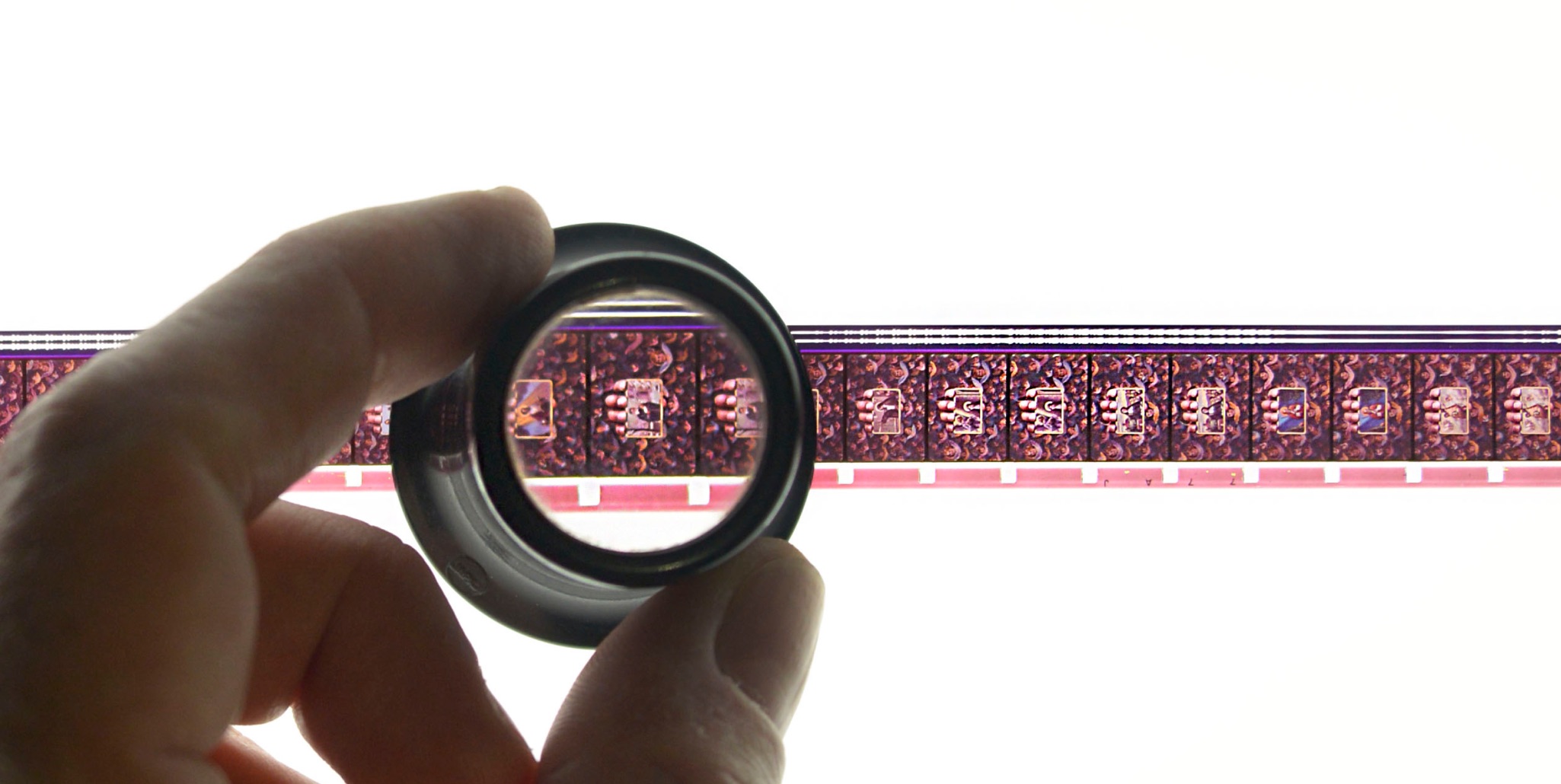 Hand holding a magnifier over a strip of 16mm film to examine individual frames.