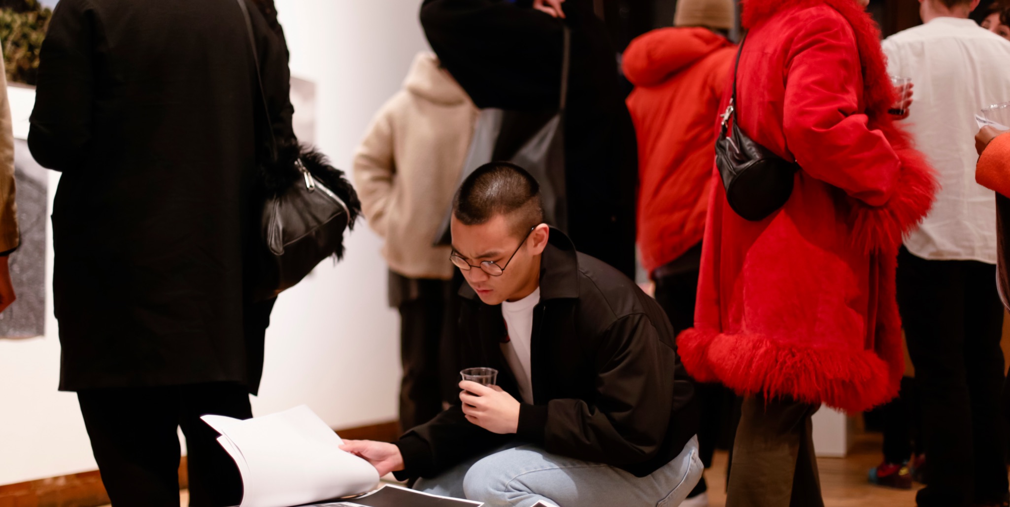 Student crouched at an exhibition, looking through a portfolio while holding a drink.