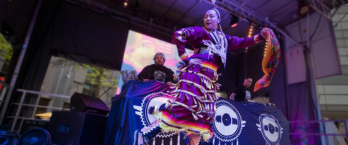 Indigenous dancer on stage with a dj performing behind her.