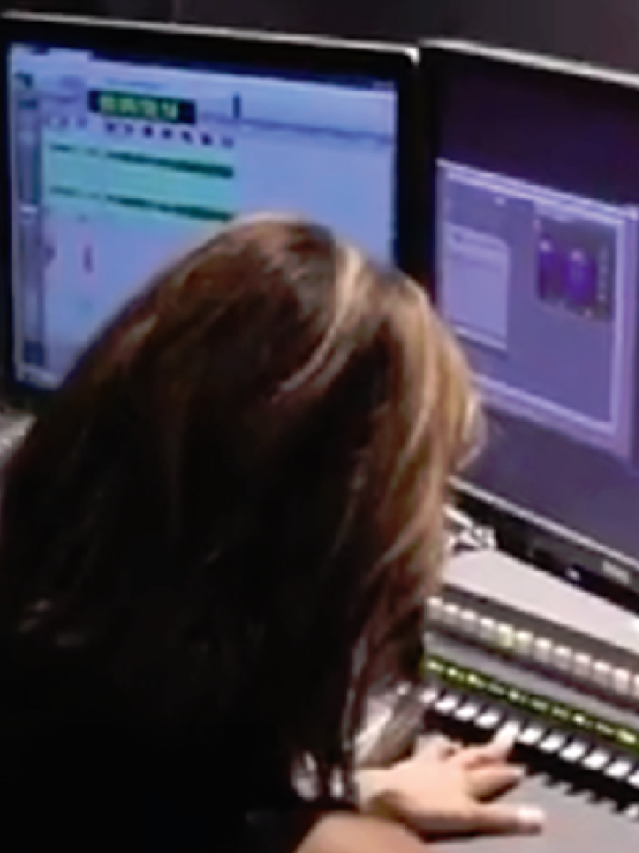 Sitting from behind, a woman is working on a mixer in front of 2 monitors showing audio visual software