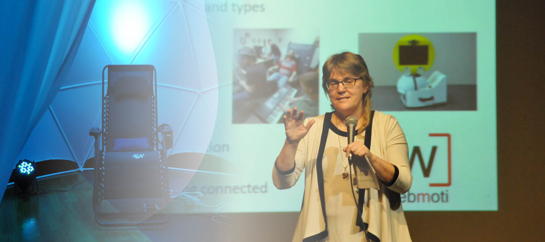 Dr. Fels holds the microphone and presents to the audience at the Sense I.T. Symposium with her slides of a black vibrating chair surrounded in blue light and a yellow circular rotating screen on a desktop with the WebMoti logo under it