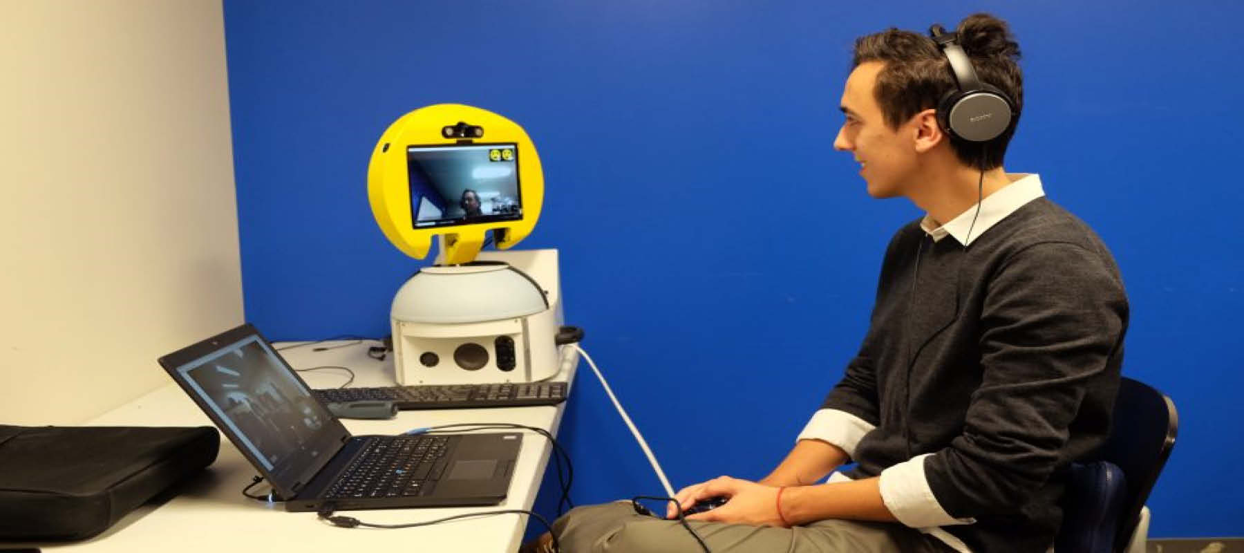 Grosu, an undergraduate student employee, sits smiling with headphones on and looking into a yellow circular, rotating screen to another person online for an accessible video conferencing application called WebMoti