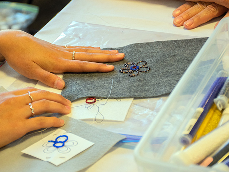 A student sewing a flower design on a piece of grey cloth.