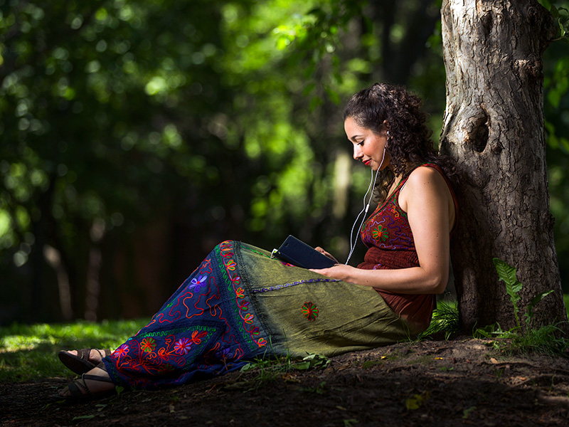 An Indigenous woman sitting by a tree while reading a book and listening to music.