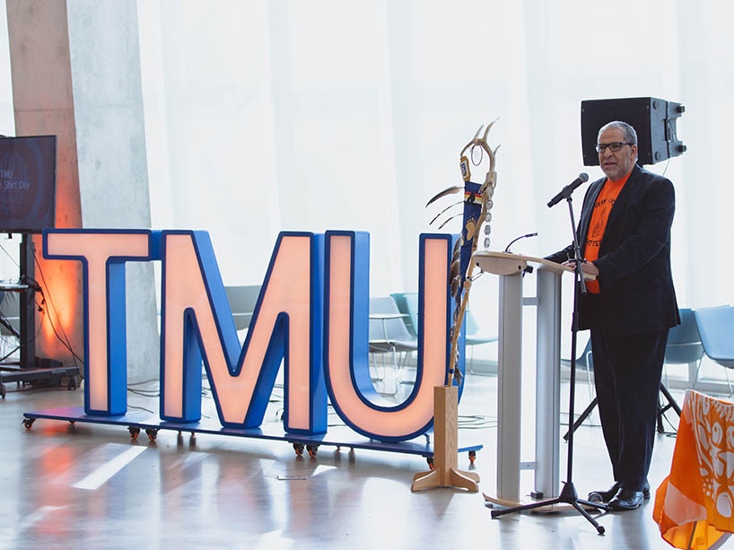 Mohamed Lachemi, TMU president, speaking at the microphone next to the Eagle Staff and the letters TMU in orange