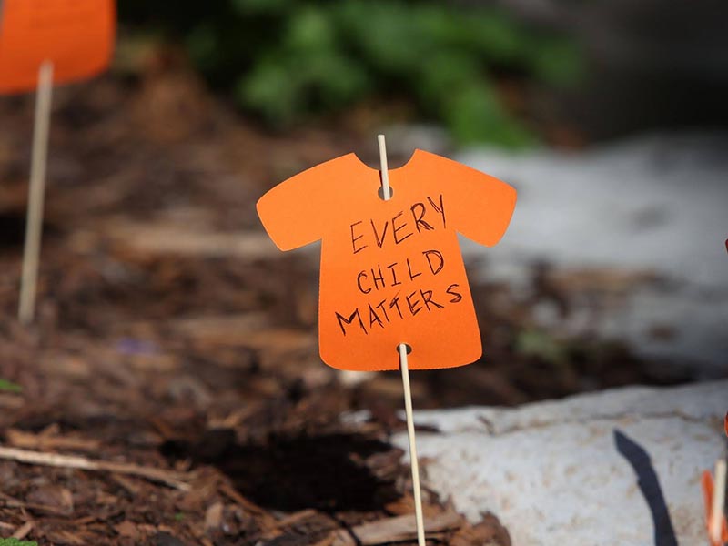 An paper cut out orange shirt, reading Every Child Matters, on a wooden stick put into the earth.