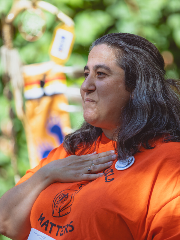 A community member in an orange shirt holds their hand to the chest while talking