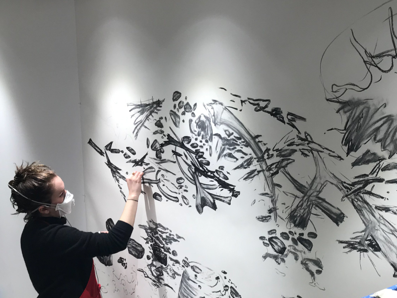 A student working meticulously on a large-scale drawing hung on the wall of the open space. Spotlights elegantly illuminate the black and white drawing.