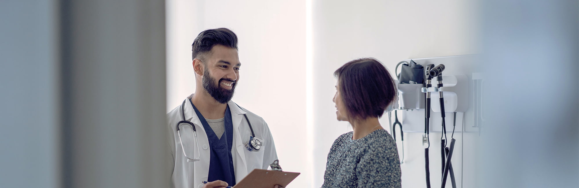 A male doctor with a beard, wearing a white lab coat and stethoscope, smiles as he talks to an older woman. He is holding a clipboard and looking at her, and she is listening to him.